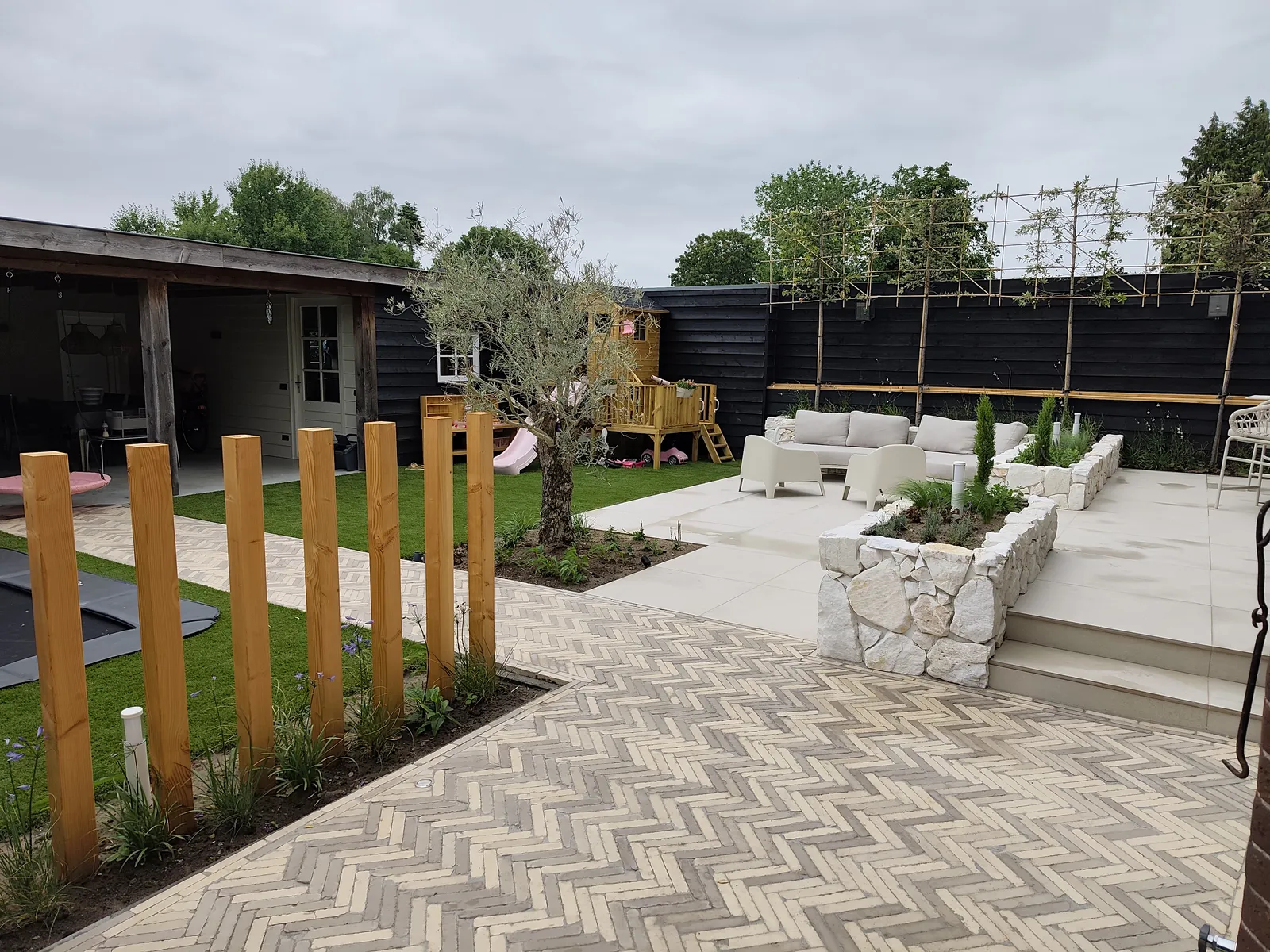 Terras van lichte keramische tegels naast een pad in visgraatverband, met verhoogde borders van natuursteen en een olijfboom