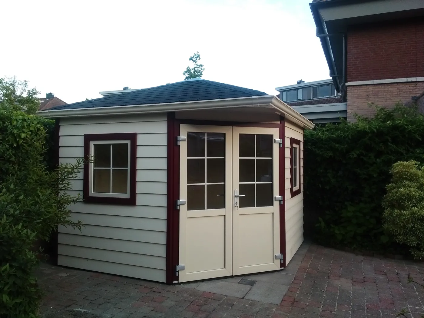 Kunststof tuinhuis met dubbele deuren en een venster met donkere omlijsting, op een bestrate ondergrond in een tuin
