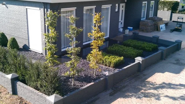 Onderhouden voortuin met geschoren buxusblokken, jonge bomen in een border van zwart grind en een muurtje van gestapelde stenen