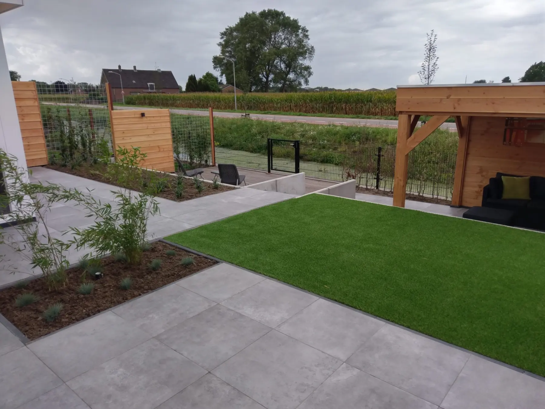 Terras van grote grijze tegels met kunstgras en een houten overkapping, met uitzicht over het land achter de tuin
