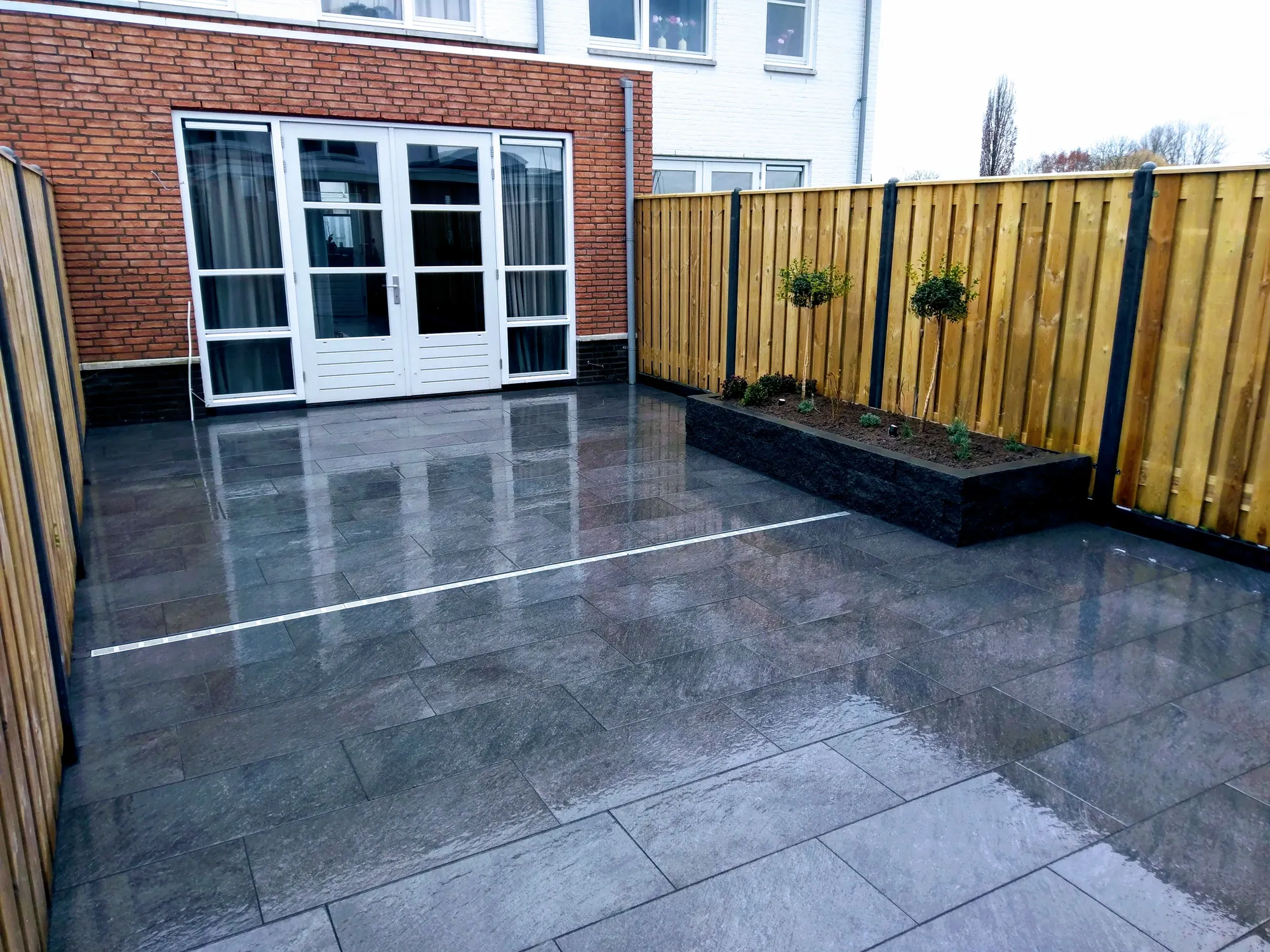 Terras van natuursteentegels tussen twee hout-betonschuttingen, met verhoogde borders langs de achtergevel