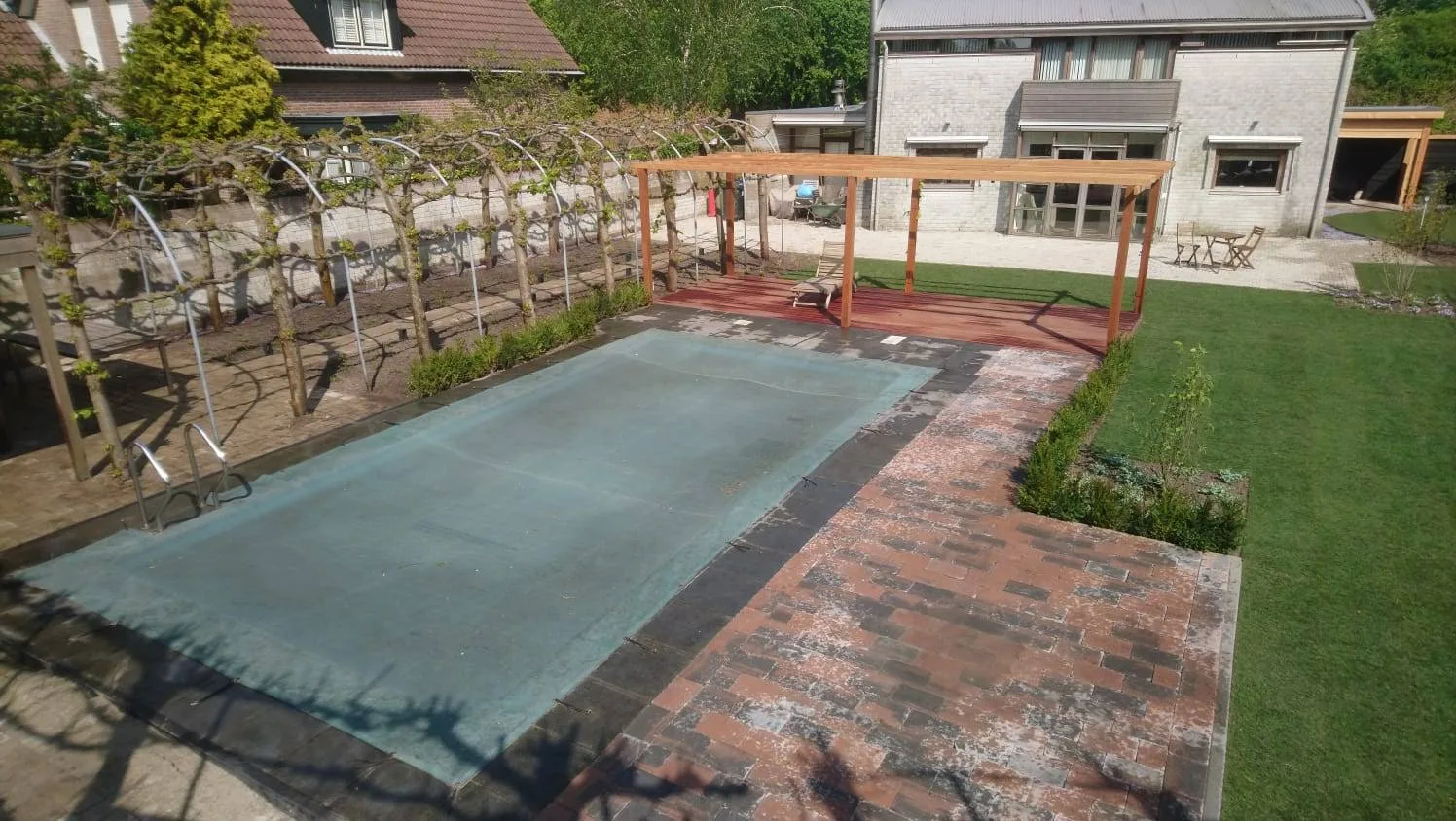 Gerenoveerde tuin van bovenaf gezien, met zwembad, leibomen langs de erfgrens, klinkerbestrating en een houten pergola