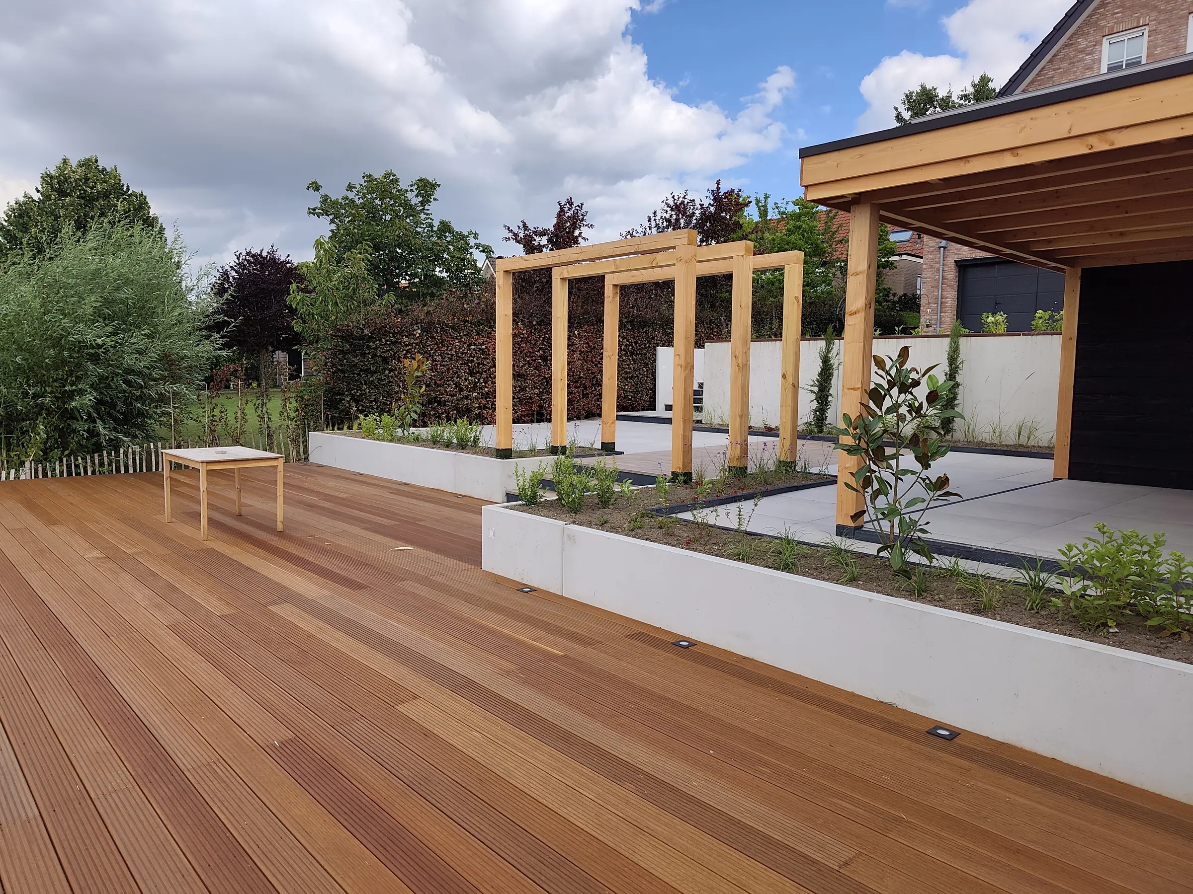 Pas aangelegde tuin met een hardhouten vlonder, betonnen plantenbakken, houten pergolaportalen, een keramisch terras en een overkapping langs een beukenhaag
